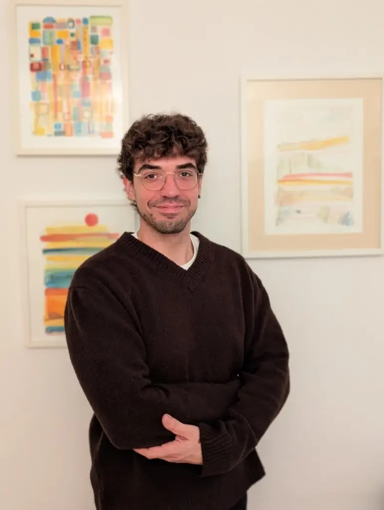 Foto d'Abel Muñoz, psicòleg a Girona, somrient amb els braços creuats davant de pintures abstractes de colors vius en una paret blanca.