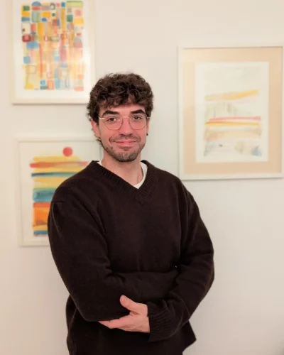 Foto d'Abel Muñoz, psicòleg a Girona, somrient amb els braços creuats davant de pintures abstractes de colors vius en una paret blanca.